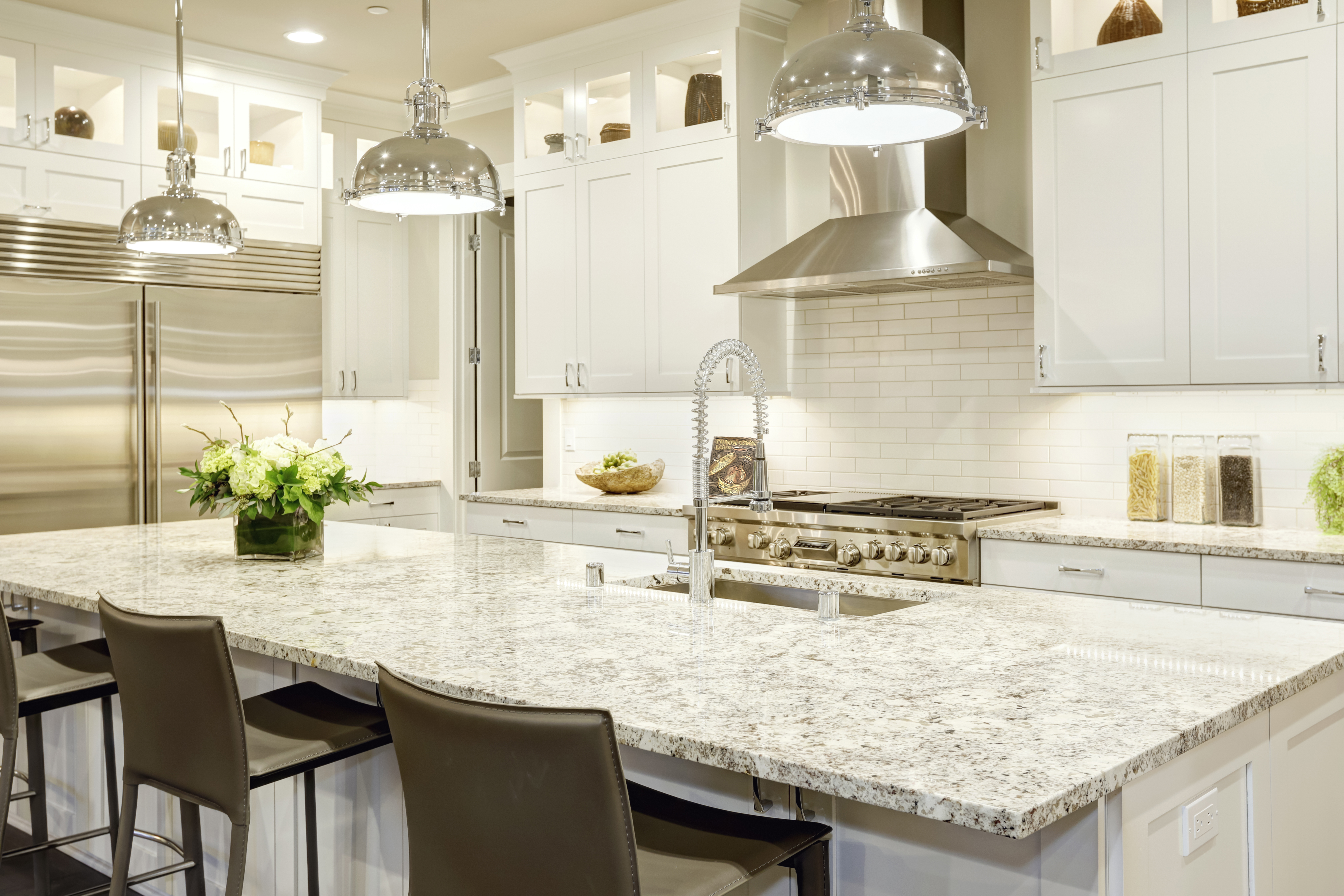 White kitchen design features large bar style kitchen island with granite countertop illuminated by modern pendant lights. Stainless steel appliances framed by white shaker cabinets